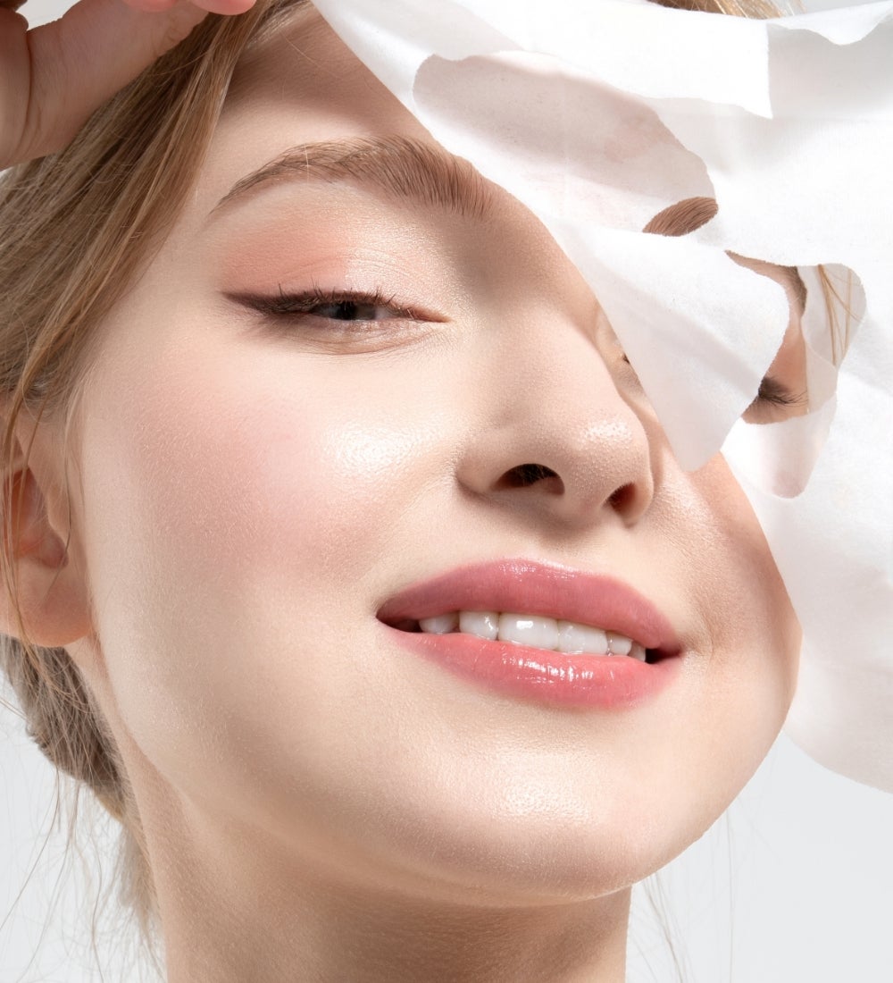 A smiling woman peeling off a Collagen Sheet Mask from her face.
